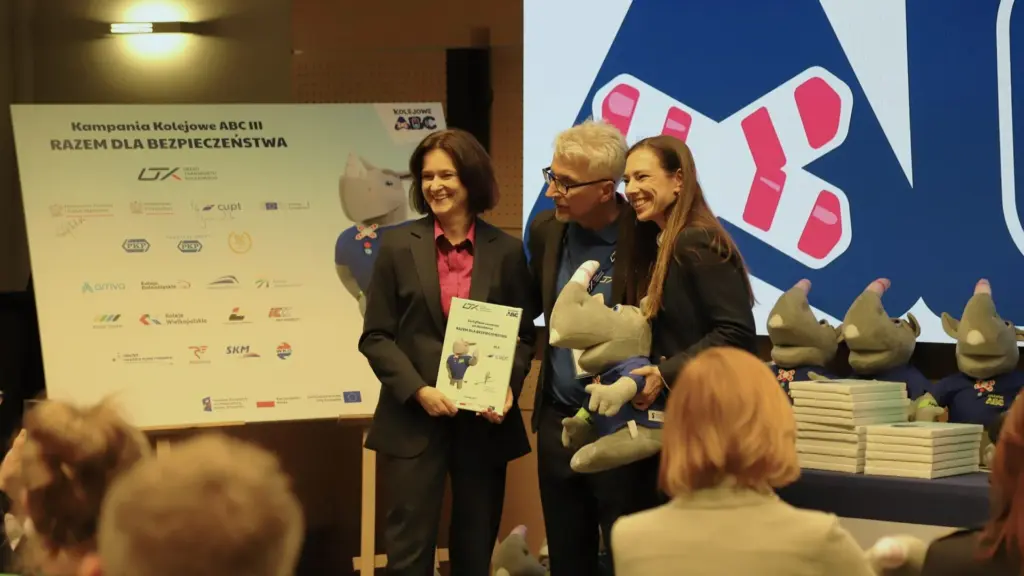 Trójka uśmiechniętych przedstawicieli na scenie podczas wydarzenia kampanii Kolejowe ABC III. Kobieta po lewej prezentuje certyfikat uznania dla CUPT, a mężczyzna obok trzyma pluszową maskotkę nosorożca Rogatka. W tle duża tablica z hasłem Razem dla bezpieczeństwa i logotypami partnerów kolejowych.