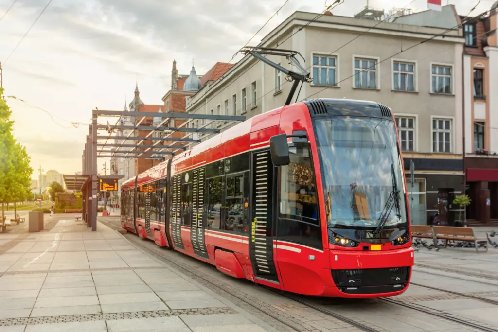 Długi, nowoczesny czerwony tramwaj stojący na peronie przystankowym obok szklanej wiaty, na tle starych miejskich kamienic. Fotografia nawiązuje do unijnych inwestycji w śląską komunikację tramwajową.