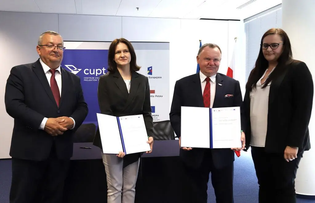 Cztery osoby (dwie kobiety i&nbsp;dwóch mężczyzn) stojące w&nbsp;rzędzie w&nbsp;sali konferencyjnej, trzymające podpisane dokumenty po&nbsp;uroczystym podpisaniu umowy w&nbsp;siedzibie CUPT.
